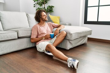 Sticker - Young hispanic man drinking coffee sitting on the floor at home.