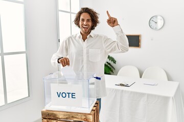 Sticker - Young hispanic man voting putting envelop in ballot box smiling amazed and surprised and pointing up with fingers and raised arms.