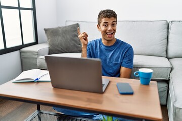 Sticker - Young handsome hispanic man using laptop sitting on the floor with a big smile on face, pointing with hand and finger to the side looking at the camera.