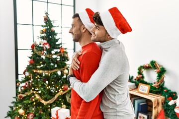 Poster - Young gay couple standing by christmas tree wearing hat looking to side, relax profile pose with natural face and confident smile.