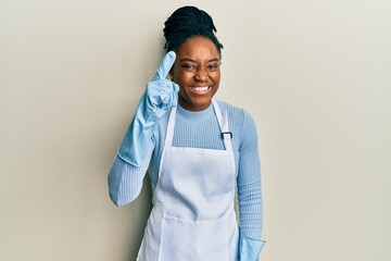 Sticker - African american woman with braided hair wearing cleaner apron and gloves pointing finger up with successful idea. exited and happy. number one.