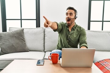 Wall Mural - Young hispanic man with beard wearing call center agent headset working from home pointing with finger surprised ahead, open mouth amazed expression, something on the front