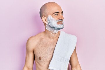 Canvas Print - Young bald man shirtless shaving beard with foam looking away to side with smile on face, natural expression. laughing confident.