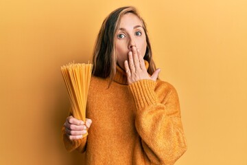 Poster - Young blonde woman holding dry spaghetti covering mouth with hand, shocked and afraid for mistake. surprised expression