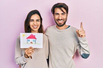 Young hispanic couple holding house draw smiling with an idea or question pointing finger with happy face, number one