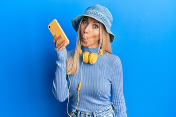 Wall Mural - Young blonde woman using smartphone wearing headphones scared and amazed with open mouth for surprise, disbelief face