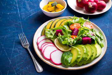 Healthy vegan salad fruits and vegetables dish with avocado, dragon fruit, radish, pumpkin, tomato, corn salad, lettuce, mizuna and lemon on blue background