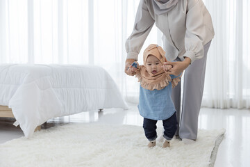 Baby girl 6 months taking her first steps and help of her mother in the bedroom. Asian family in hijab clothing , motherhood, parenting, and child care