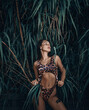 Gorgeous sexy excited brunette woman savage with slim athletic body in fashion leopard print, pattern bikini stands at tropical tree leaves, plants looking aside. Summer vibes