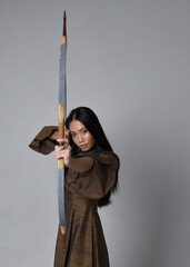 Wall Mural - Close up  portrait of beautiful young asian woman with long hair wearing medieval fantasy gown. Graceful pose holding a long bow and arrow,  isolated on studio background.