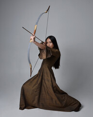 Wall Mural - Close up  portrait of beautiful young asian woman with long hair wearing medieval fantasy gown. Graceful pose holding a long bow and arrow,  isolated on studio background.