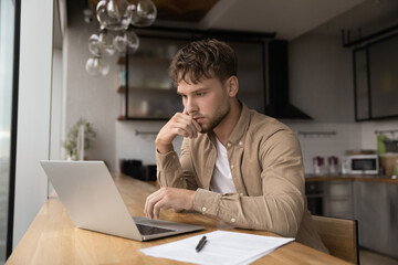 Thoughtful serious young 25s man student freelancer working or studying use laptop looks at device screen thinking solving business remotely, doing research sit alone in modern home or office kitchen