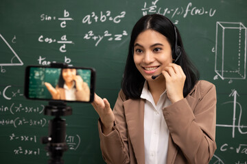 Wall Mural - Young asian teacher woman standing looking at camera and video conference with student. Female teacher training the mathematics in classroom from live stream with smartphone online course.