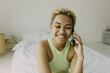 © shurkin_son - People, technology, communication and rest concept. African ethnicity woman with curly dyed white short hair laughing with eyes closed while talking on phone with bestie sitting at white bed