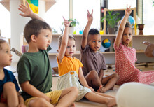 Group Of Small Nursery School Children Sitting On Floor Indoors In Classroom, Raising Hands.