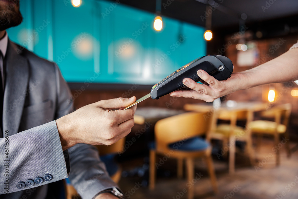 Hands holding a payment terminal. A man elegantly dressed in a business suit puts a credit or debit card in an online payment terminal held by a feminine hand. Paying bills for a meal at a restaurant - obrazy, fototapety, plakaty 