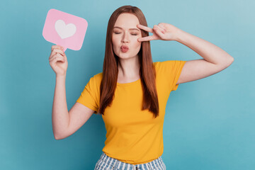 Portrait of romantic lady hold heart card like button show v-sign cover eye close eyes send air kiss on blue background