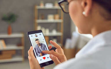 Woman looking for romantic relationship. Single lady holding smartphone and giving like to photo of young man on online dating mobile app or website. Closeup display shot, close up view over shoulder