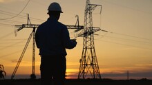Silhouette Power Engineer In White Helmet Checks Power Line Using Computer On Tablet, Remote Control Of Power System. High Voltage Electrical Lines At Sunset. Distribution And Supply Of Electricity