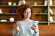 © Vadim Pastuh - Satisfied smiling young woman with curly hairstyle sitting on the couch, holding credit card and smartphone, paying in the Internet, making order, charming red-haired girl enjoys shopping from home