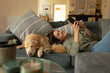 © Wavebreak Media - Smiling caucasian woman in living room, lying on sofa with her pet dog, using smartphone