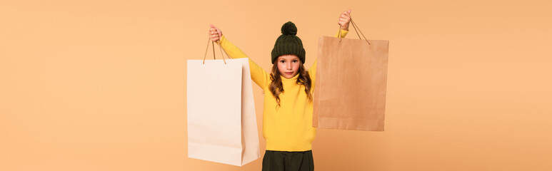 Wall Mural - fashionable kid in yellow turtleneck holding shopping bags in raised hands isolated on beige, banner