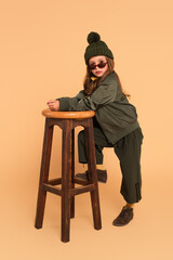 Wall Mural - fashionable girl in autumn clothes looking at camera over sunglasses near high stool on beige