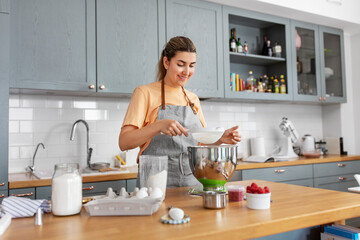 Wall Mural - culinary, baking and people concept - happy smiling young woman cooking food on kitchen at home and sifting flour through sieve