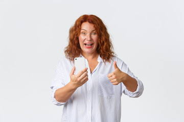 Technology and communcation concept. Amused and pleased redhead middle-aged lady recommend mobile application, holding smartphone and showing thumbs-up in approval, white background