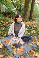 Cute girl in glasses, brown sweater, black jeans and beige scarf sitting on plaid in park and drink coffee or tea from white polka dot cup. Beverage for cold autumn days. Fallen maple leaves around.