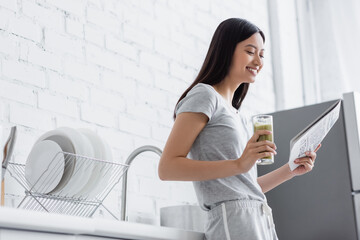 joyful asian woman with glass of fresh smoothie reading newspaper in kitchen.