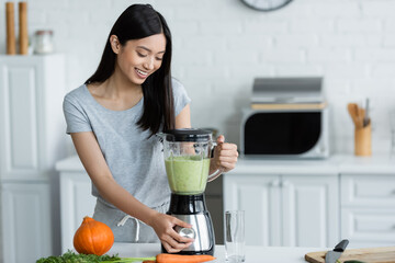 Wall Mural - smiling asian woman preparing fresh vegetable smoothie in blender near ripe pumpkin on table.