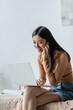 © LIGHTFIELD STUDIOS - smiling asian woman talking on smartphone near laptop in bedroom.