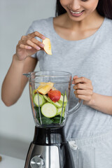 Wall Mural - partial view of smiling asian woman adding apple and zucchini into electric blender.