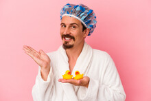 Young Caucasian Man Going To The Shower With Rubber Ducks Isolated On Pink Background Showing A Copy Space On A Palm And Holding Another Hand On Waist.