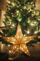 Wall Mural - Christmas star on background of christmas tree in lights in evening room. Big paper star garland glowing in festive decorated scandinavian room. Atmospheric christmas eve. Happy holidays