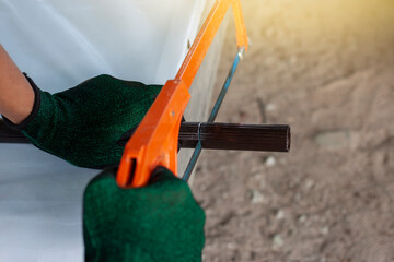 Hands of a construction worker wearing gloves is using a hacksaw to cut a piece of aluminum pipe.