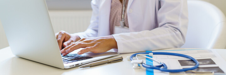 Cropped photo of female doctor in white uniform working on laptop computer in hospital, sitting at desk with stetoscope and ultrasound results. Medical panoramic banner for website header