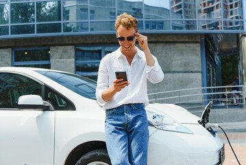 The guy sat down on the hood of the car. His car is charging at the charging station. A man looks at the smartphone screen and smiles.