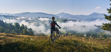 Man Riding Bicycle On Grassy Hill And Looking At Beautiful Misty Mountains. Male Bicyclist Enjoying Panoramic View Of Majestic Mountains During Bicycle Ride. Concept Of Sport, Bicycling And Nature.