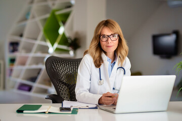 Smiling middle age female doctor sitting at desk and working on laptop at the doctor’s office. 