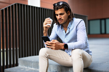 Young man sitting on the stairs drinking coffee. Handsome businessman talking to the phone while drinking coffee..