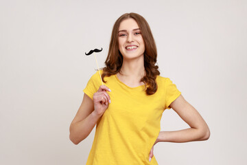 Wall Mural - Portrait of funny playful wavy haired teen girl in urban style T-shirt keeping fake paper mustache and looking at camera with happy expression. Indoor studio shot isolated on gray background.