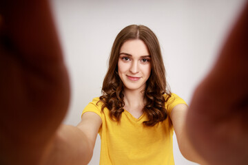 Wall Mural - Portrait of beautiful pleasant looking teenager girl in yellow casual T-shirt taking selfie, looking at camera with positive emotion POV. Indoor studio shot isolated on gray background.