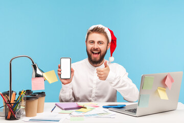 Wall Mural - happy positive man in santa claus hat and white shirt showing thumbs up gesture holding empty displa