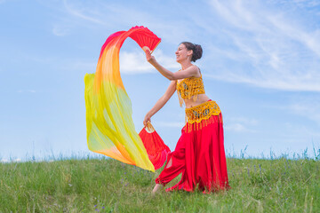 Wall Mural - Young woman in a bright national costume is dancing an oriental dance with fans on a green meadow against a blue summer sky. Pleasant pastime.