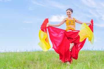 Wall Mural - Young woman in a bright national costume is dancing an oriental dance with fans on a green meadow against a blue summer sky. Pleasant pastime.