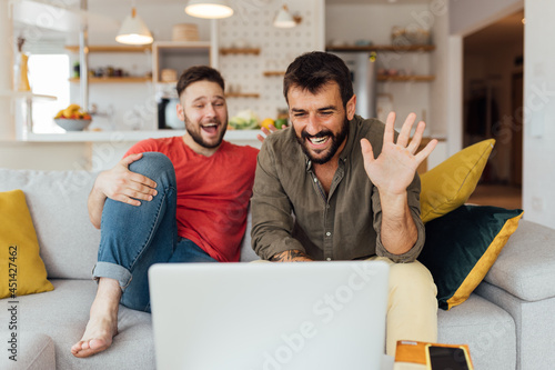 Young happy Caucasian gay couple in love sitting in their cozy living room and having video call on the laptop with their friends. Since it\'s a corona virus outbreak we have to stay safe at home.