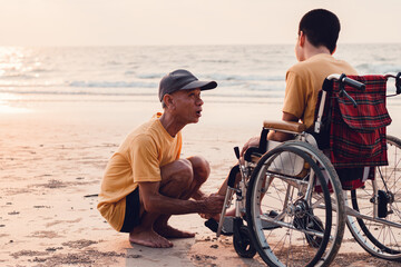 Wall Mural - Happy disabled teenage boy on wheelchair playing and relaxing with parent, Activity outdoors with father on the beach background, People having fun and diverse family concept.