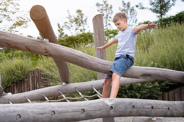 Wall Mural - Cute blond boy the boy sitting on a wooden beam on playground in public park.